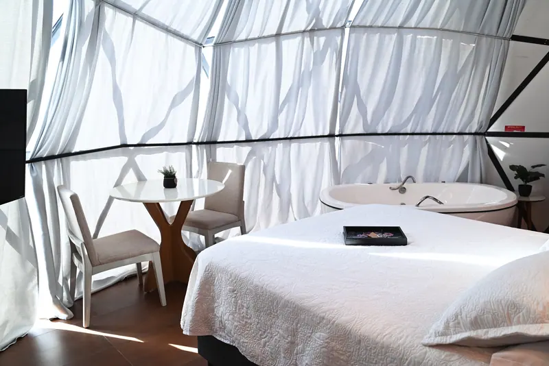 Interior do domo com cama de roupa branca, mesa redonda e banheira